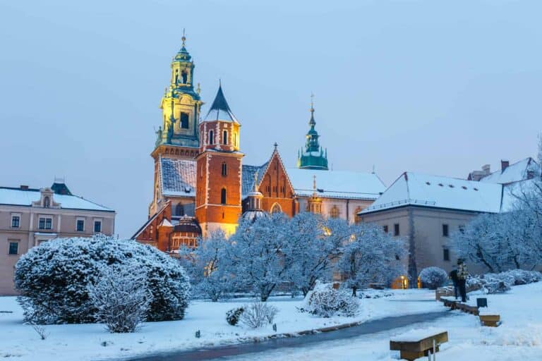 A historic brick cathedral with green spires stands amid snow-covered trees and buildings on a winter evening in Krak&oacute;w&mdash;a magical sight among the best things to do in December.