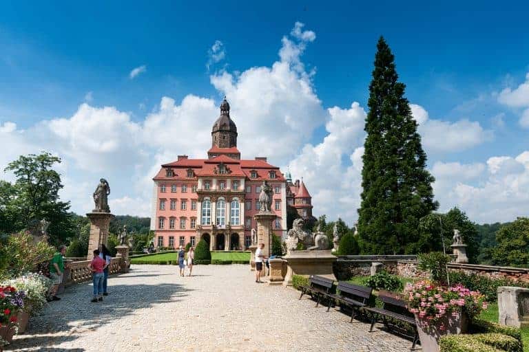 People walk along a stone pathway leading to a large, historic pink castle&mdash;one of the best places to visit in Dolnośląskie, Poland&mdash;surrounded by statues, benches, plants, and a tall evergreen tree under a blue sky.