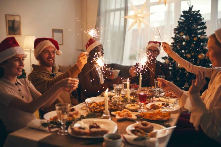 Five people wearing Santa hats sit around a festive table with food, holding sparklers and celebrating beside a decorated Christmas tree, embracing Polish New Year's Traditions.