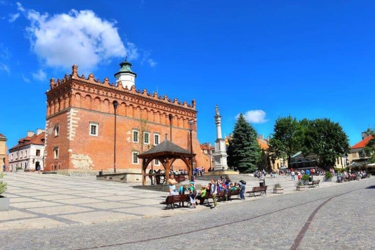 A historic brick building with a clock tower stands in a sunny town square in Swietokrzyskie, Poland&mdash;one of the best places to visit&mdash;surrounded by people on benches and strolling past trees and smaller buildings in the background.