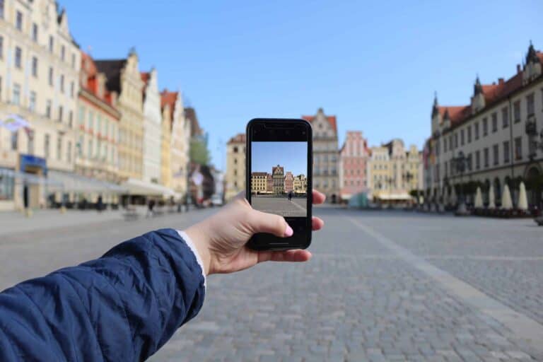A person holds a smartphone, taking a photo of colorful buildings in an empty European city square on a clear day.