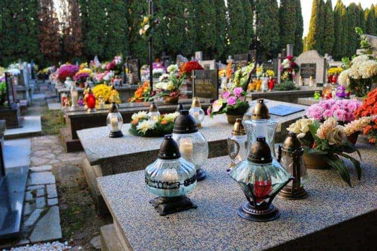 Gravestones in a cemetery decorated with colorful flowers and glass lanterns, surrounded by tall evergreen trees, capture the serene beauty of autumn in Poland.