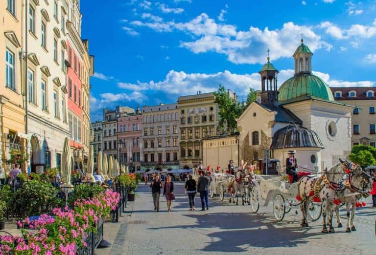 First time visitors to Krakow stroll along a cobblestone street with horse-drawn carriages, colorful buildings, and a historic church under a bright blue sky.