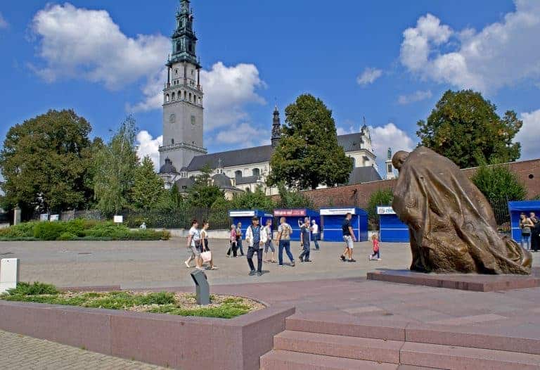 People walk near a large bronze statue and a church with a tall bell tower, surrounded by trees under a partly cloudy sky&mdash;a scene typical of top sites on a Poland pilgrimage.