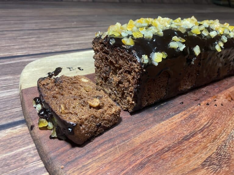 Loaf of chocolate gingerbread cake with nuts and candied fruit on top, partially sliced, on a wooden cutting board.