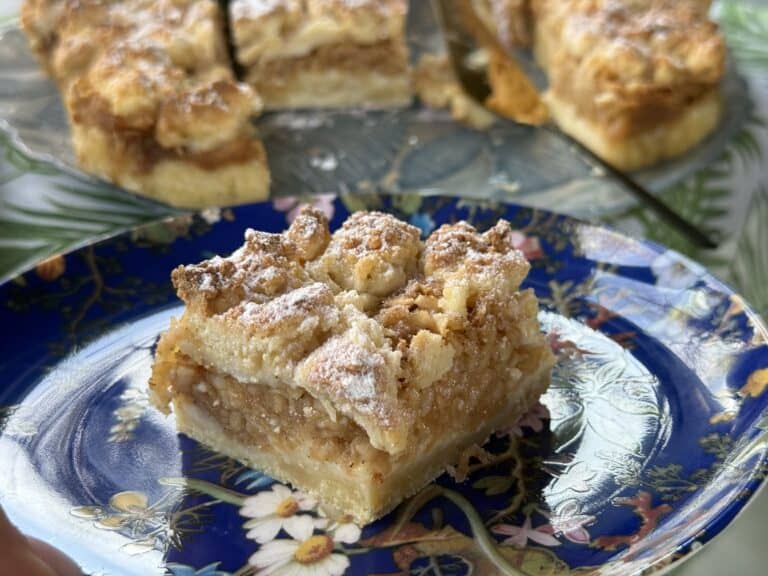 A slice of Polish apple cake, known as Jablecznik, rests on a floral-patterned blue plate, with more delectable pieces in the background on another plate.