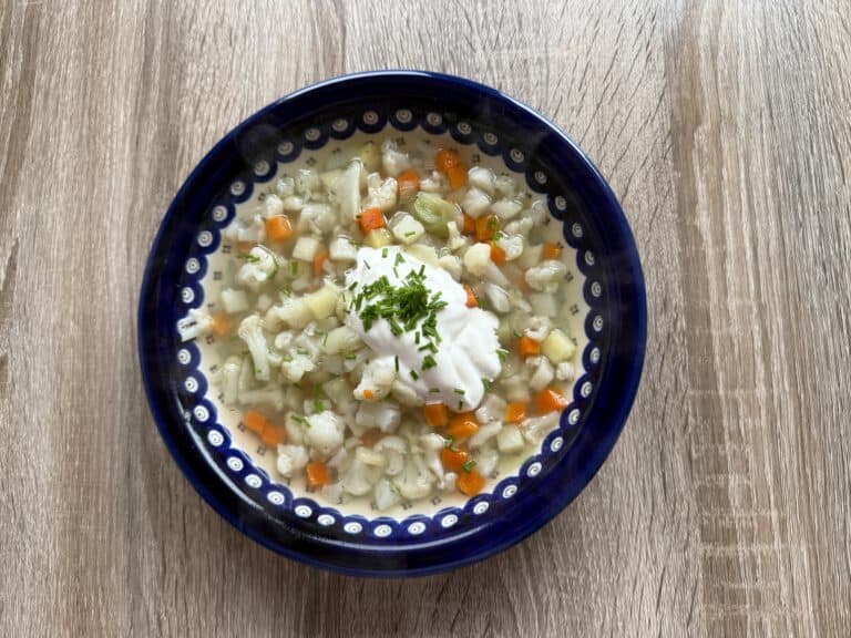 A bowl of creamy Polish Cauliflower Soup with diced carrots, potatoes, and herbs, topped with a dollop of sour cream and chopped chives, on a wooden table.