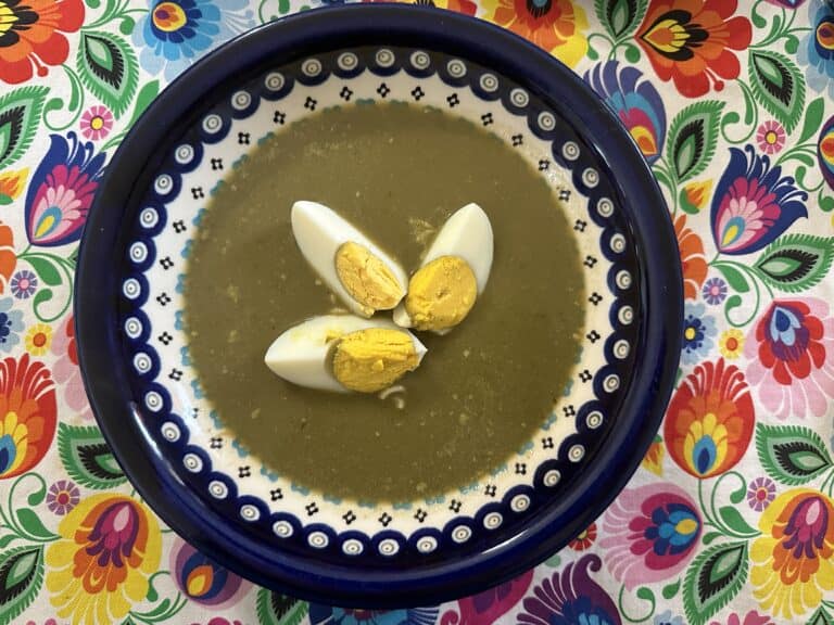 A bowl of Polish sorrel soup with three slices of hard-boiled egg, elegantly served in a blue and white patterned dish, rests on a colorful floral tablecloth.