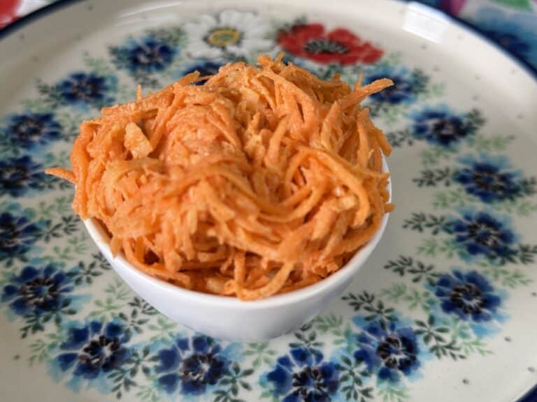 A small white bowl filled with shredded orange carrot, inspired by a Polish carrot salad recipe, sits on a decorative plate with blue and red floral patterns.