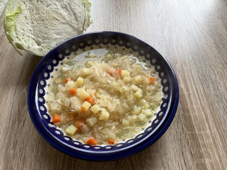 A bowl of Vegetarian Kapusniak, filled with diced potatoes and carrots, sits on a wooden table next to a half head of cabbage.