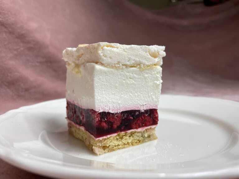 A slice of malinowa chmurka dessert with a white top, red fruit middle, and a beige base sits elegantly on a white plate.