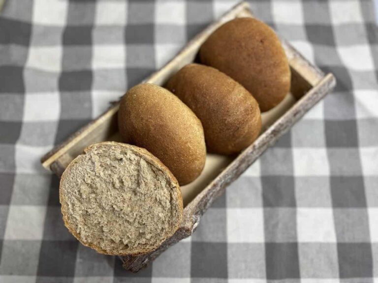 Four round, Polish whole bread rolls are placed in a wooden tray on a checkered tablecloth. One roll is sliced in half, revealing its textured interior.