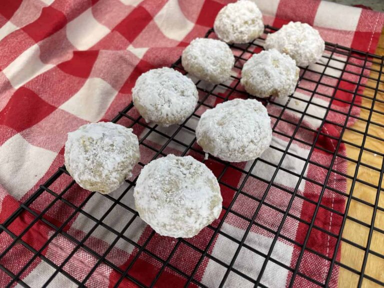 Eight Polish snowball cookies, covered in powdered sugar, are cooling on a black wire rack placed on a red and white checkered cloth.