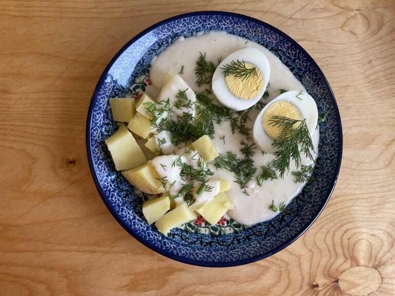 A plate of Polish Eggs in Horseradish Sauce featuring potatoes and hard-boiled eggs topped with a creamy white sauce, garnished with fresh dill, served on a blue-patterned dish on a wooden table.