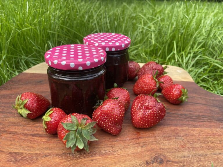 Two jars of dżem truskawkowy with pink polka-dot lids are placed on a wooden board, surrounded by fresh strawberries, with green grass in the background.