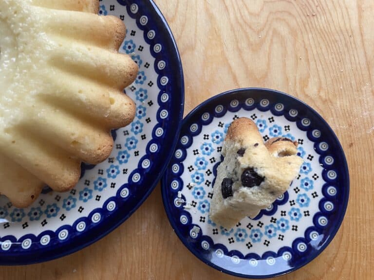 A whole bundt cake, reminiscent of a Polish Babka Z Jagodami, sits on a blue patterned plate with a slice showcasing dark fruit pieces on a smaller similar plate. Both are elegantly placed on a wooden surface, inviting you to savor the delightful blueberry babka flavor.