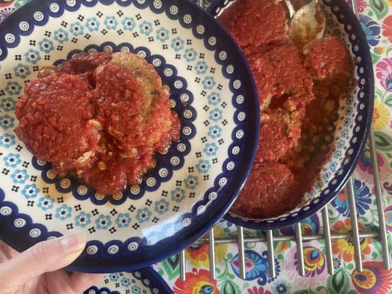 A hand holds a plate with stuffed artichokes topped with tomato sauce, reminiscent of a Gołąbki Casserole, above a larger dish containing more servings on a colorful tablecloth. Perfect for those seeking an unstuffed cabbage rolls vibe in a quick recipe format.