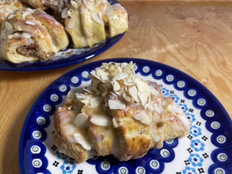 A pastry with icing and sliced almonds sits enticingly on a blue patterned plate, reminiscent of traditional Rogale Świętomarcińskie. In the background, another Polish Saint Martin's Croissant awaits, showcasing the charm of this cherished recipe.