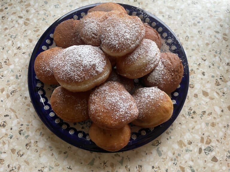 A plate of sugar-dusted vegan pączki on a patterned blue dish rests elegantly on a speckled countertop.