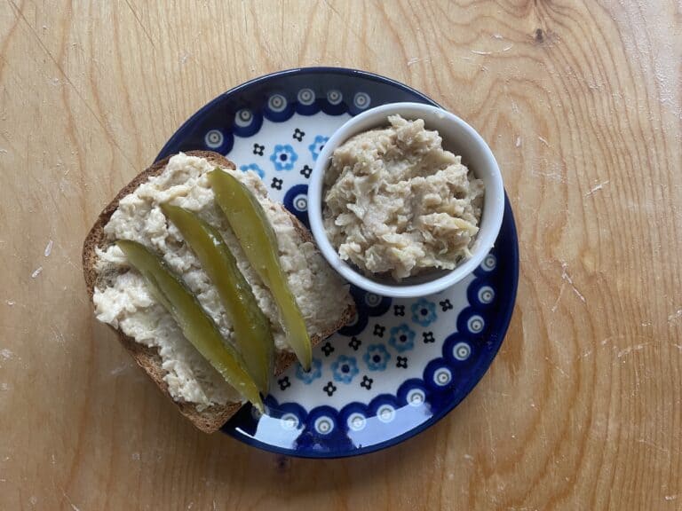 Bread topped with a delightful Polish Vegan lard spread and pickles rests on a patterned plate, accompanied by a small bowl of Smalec Wegański.