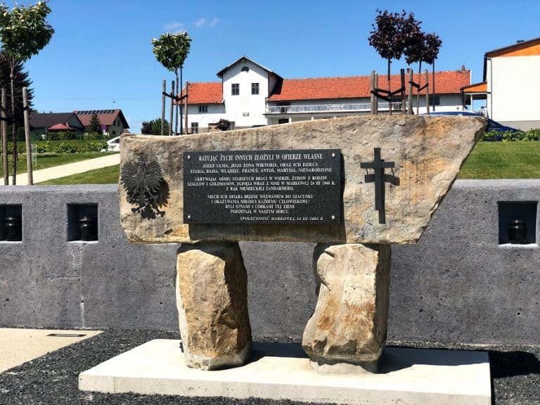 A stone memorial monument with a plaque, Christian cross, and coat of arms stands outdoors in front of houses and trees&mdash;a notable site in Lancut, Poland for any travel guide or those seeking insider tips.