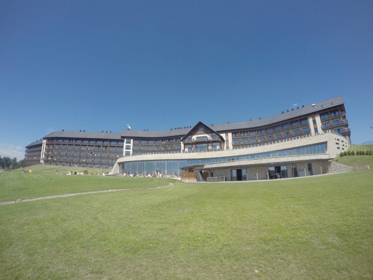 A large modern hotel with multiple stories and balconies is situated on a grassy lawn under a clear blue sky, offering an ideal retreat for exploring the Bieszczady Mountains in Poland&mdash;an insider&rsquo;s guide to comfort and adventure.