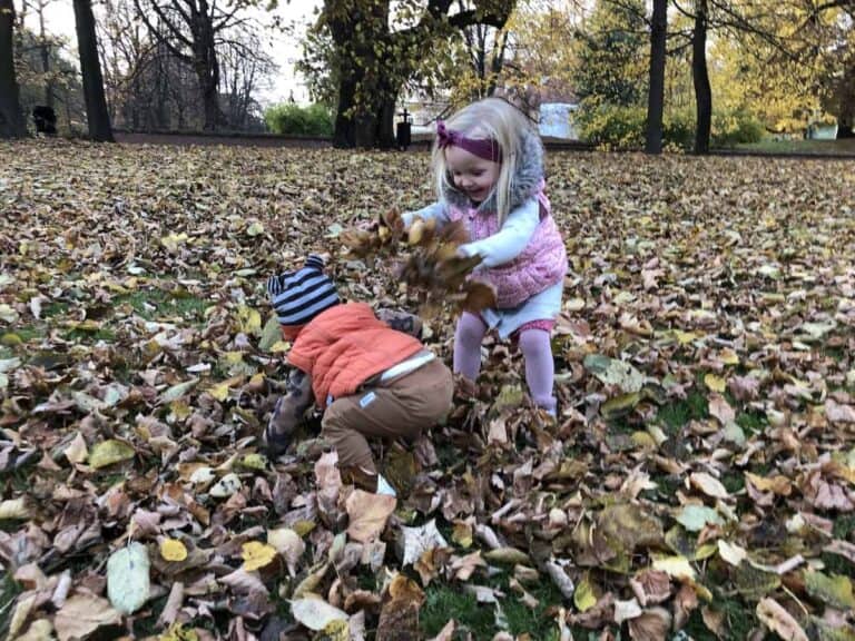 Two young children play in a park, surrounded by fallen leaves. One child stands, smiling, and tosses leaves onto the other, capturing the joyful spirit of Autumn in Poland.