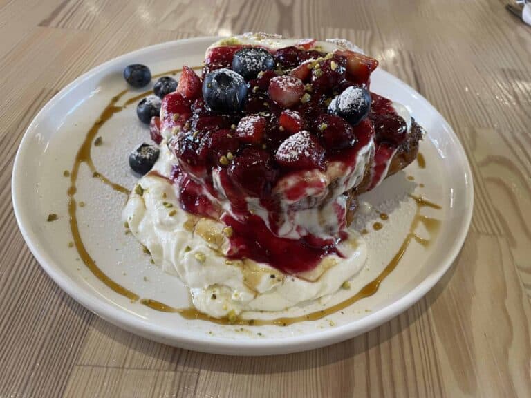 A plate with stacked pancakes topped with whipped cream, berry compote, blueberries, strawberries, powdered sugar, and a drizzle of caramel sauce.