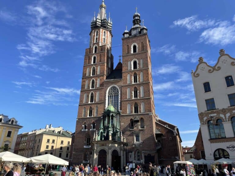 A large brick church with two tall towers and a central entrance stands in a busy square under a blue sky, surrounded by historic buildings and people&mdash;one of the top things to see in Poland on any 5 days itinerary.