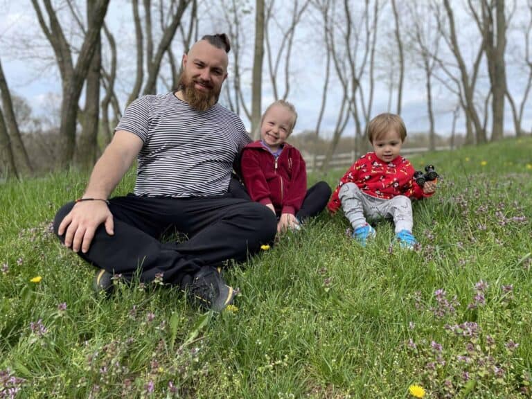 An adult and two young children sit on the grass in a park with trees in the background, all facing the camera and smiling.