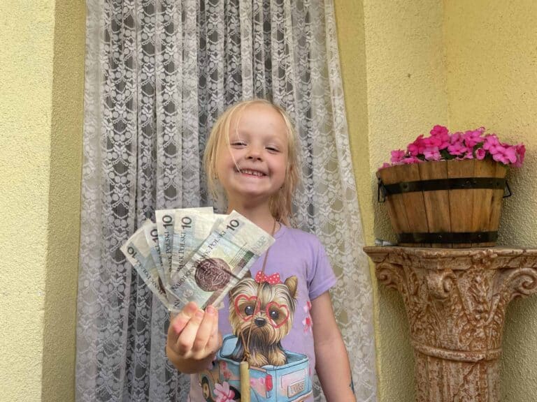 Young girl smiling and holding several ten-pound British banknotes, standing in front of a lace curtain and a flower pot&mdash;perhaps saving up after discovering how cheap travel in Poland can be.