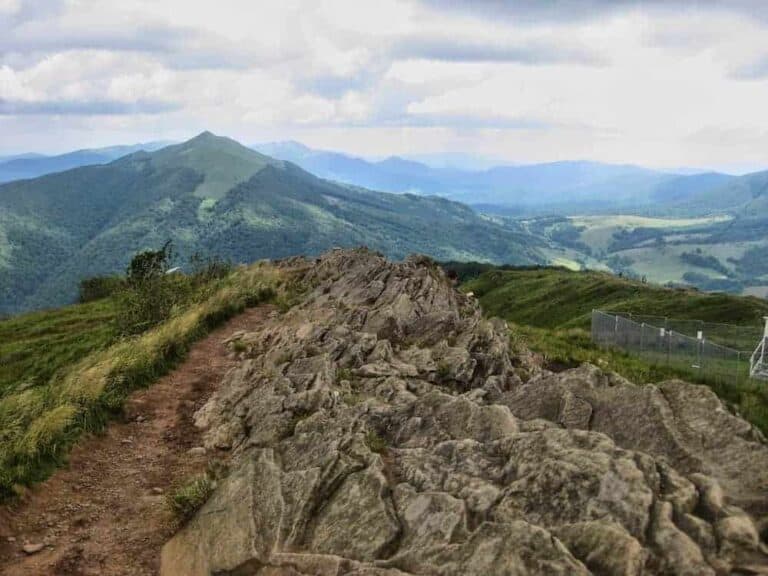 A rocky trail runs along a ridge with grassy hills and distant mountains under a cloudy sky. A metal fence is visible on the right side.