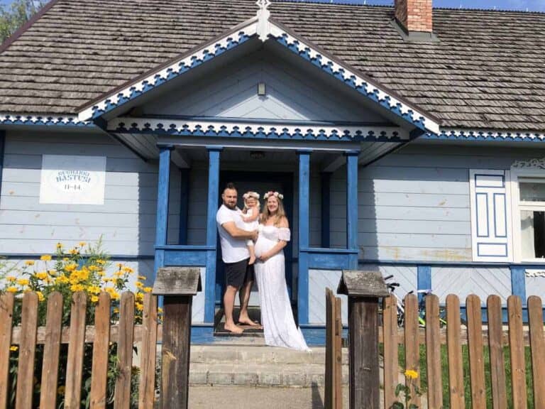 A man, woman, and child stand together in front of a blue wooden house with a picket fence and yellow flowers, capturing the charm of Poland summer.