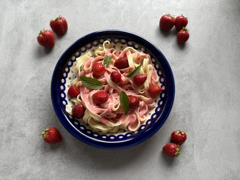 A bowl of tagliatelle pasta with a creamy pink sauce, inspired by makaron z truskawkami, is garnished with fresh strawberries and mint leaves, sitting on a grey surface and surrounded by whole strawberries.