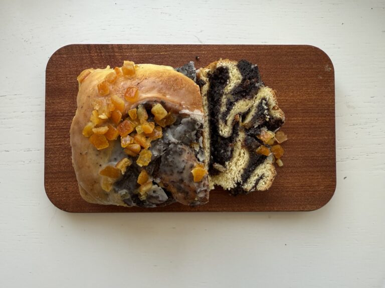 A loaf of Makowiec Zwijany, or poppy seed swirl bread, topped with diced candied orange peel and partially sliced, rests on a wooden board.