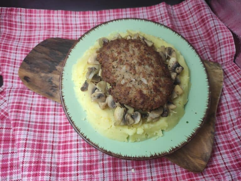 A plate of mashed potatoes topped with saut&eacute;ed mushrooms and a breaded meat patty, reminiscent of a Polish Bryzol Recipe, served on a wooden board placed over a red and white checkered cloth.