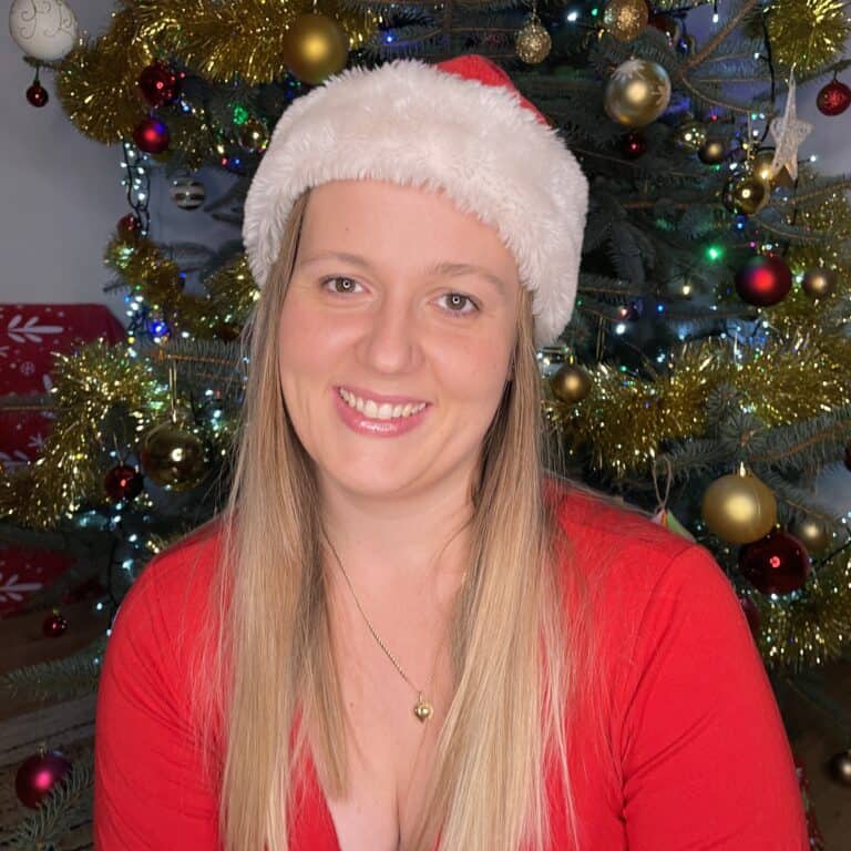 Person in a Santa hat and red outfit smiling in front of a decorated Christmas tree with lights and ornaments.