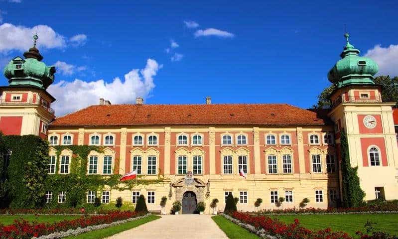 A large, ornate palace with a red-tiled roof, two green-domed towers, and manicured gardens under a blue sky—one of the best places to visit when you travel through Podkarpackie, Poland.