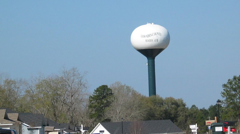 The water tower for Lowndes County, which implemented data analytics for its water system, sprouts into the sky behind house roofs and some trees.