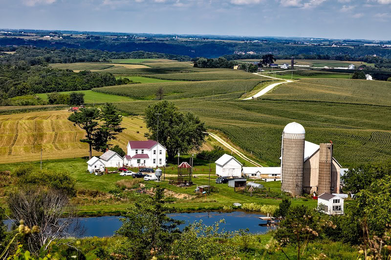 Iowa farmstead next to a manmade lake with rolling hills and a blue sky as a backdrop.