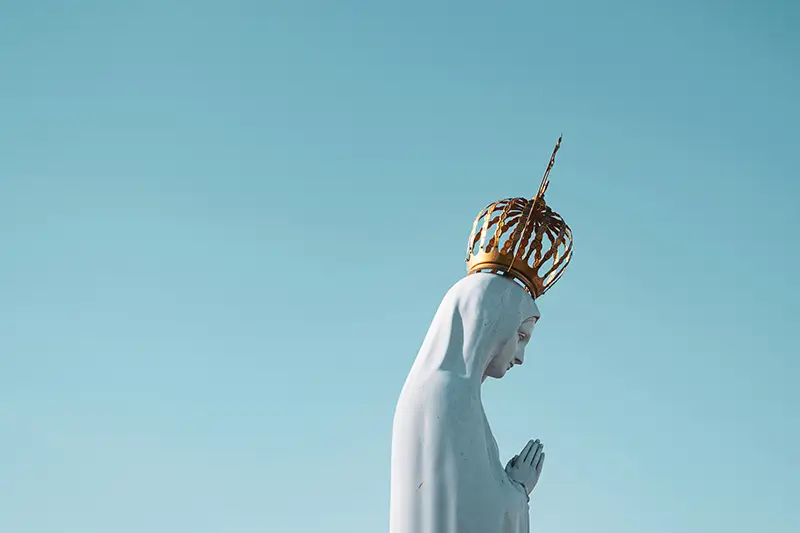 Estatua de la Virgen Mar&iacute;a con corona dorada sobre un fondo azul claro, simbolizando su realeza.