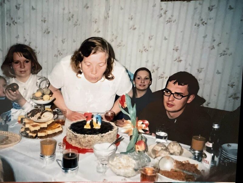 A Polish woman blows out candles on a cake numbered 70 at a dining table, surrounded by fellow foodies enjoying various foods and drinks.