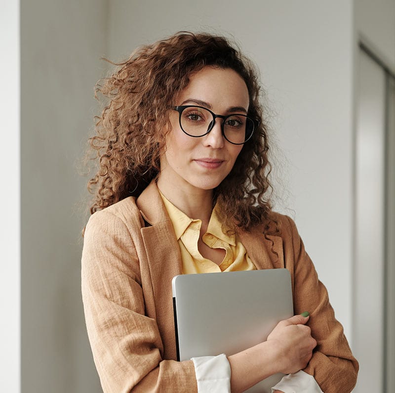 Confident businesswoman with curly hair and glasses holding a laptop, wearing a tan blazer and yellow blouse.