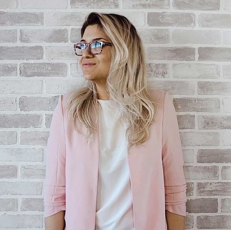 Smiling woman with long blonde hair and glasses, wearing a pink blazer and white shirt, standing against a light brick wall.