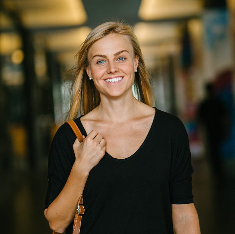 Smiling woman with blonde hair wearing a black top and carrying a brown shoulder bag, standing indoors with a blurred background.