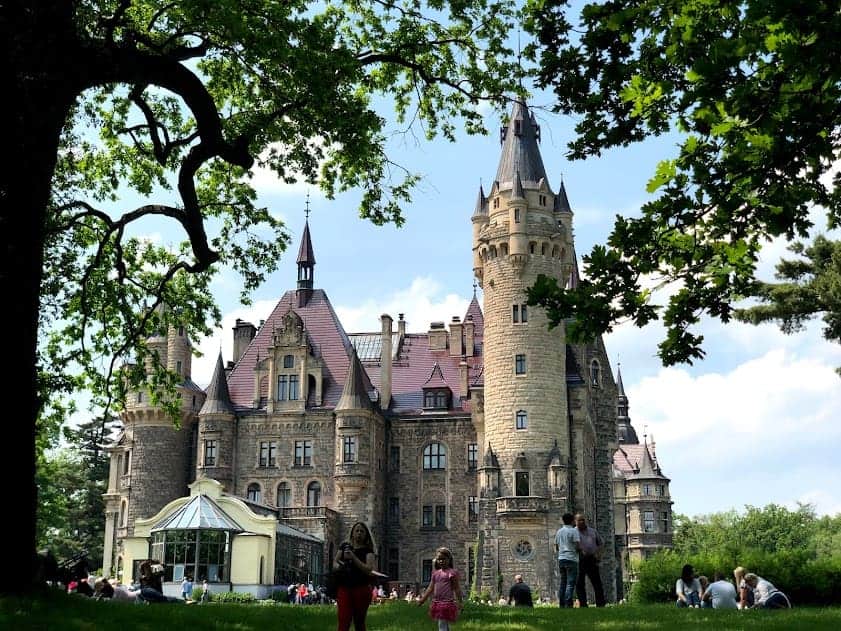 Large stone castle with turrets and red roofs, surrounded by people relaxing on a grassy lawn under leafy trees on a sunny day&mdash;a must-see castle for anyone interested in Poland tourism.