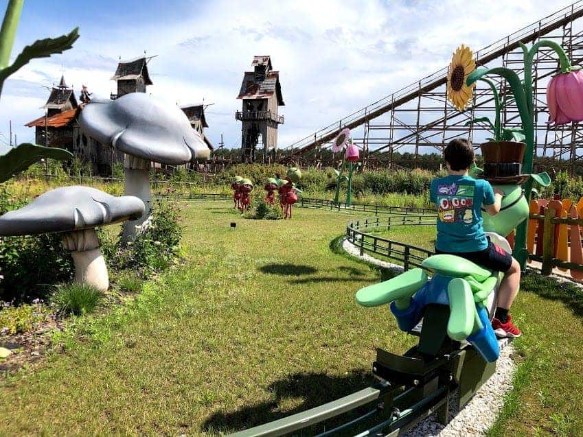 A child rides a green insect-themed ride on a track at Majaland Amusement Park Kownaty, surrounded by oversized mushroom and flower decorations. A wooden roller coaster stands tall in the background.
