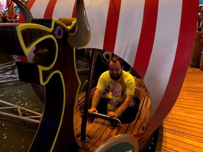 An adult and a young child sit together on a Viking-themed boat ride with red and white striped sails at Majaland Kownaty amusement park.