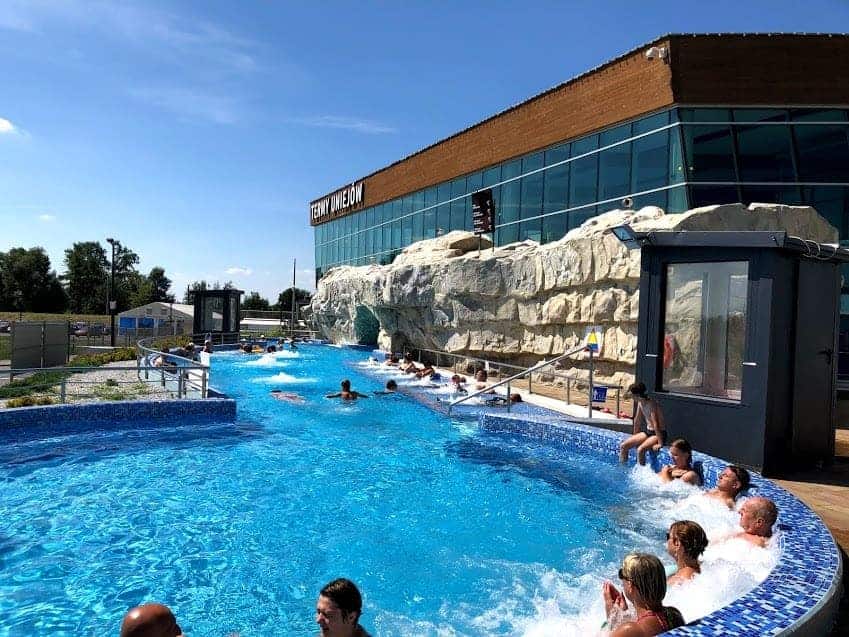 People relax in an outdoor lazy river pool next to a modern building with rock-like features under a sunny sky&mdash;a great spot among the top places to visit in Ł&oacute;dź Voivodeship.