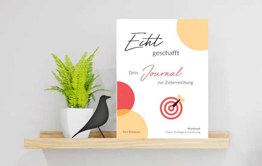 „Buch ‚Echt geschafft – Dein Journal zur Zielerreichung‘ von Tara Riedman steht auf einem hellen Holzregal neben einer grünen Pflanze und einer stilisierten schwarzen Vogel-Figur. Das Cover zeigt bunte Kreise und eine Zielscheibe mit Pfeil, darunter der Hinweis: Workbook für Vision, Strategie & Umsetzung.